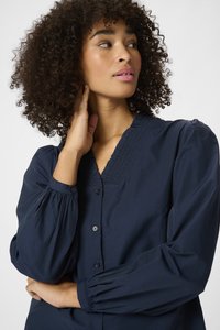 Chemise boutonnière bleu marine avec un décolleté en V plissé, des manches bouffantes et des détails texturés. Tissu lisse avec une coupe décontractée.