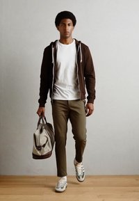 Sudadera con cremallera marrón sobre una camiseta blanca, pantalones verdes oliva y zapatillas gris y verde. Llevando una bolsa de deporte gris con acentos marrones.
