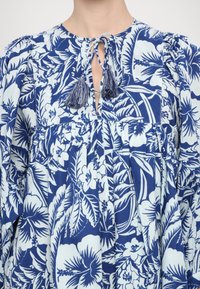 Blauwe bloemenjurk met een ontspannen pasvorm, voorzien van een split-neklijn die is vastgebonden met franjes, en geplooide details in een lichtgewicht stof.