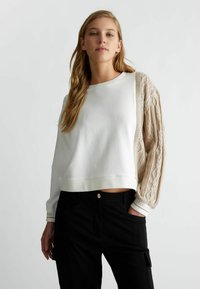 Felpa bianca corta con manica in maglia a treccia beige testurizzata, dotata di scollo rotondo e orlo a coste, abbinata a pantaloni neri.