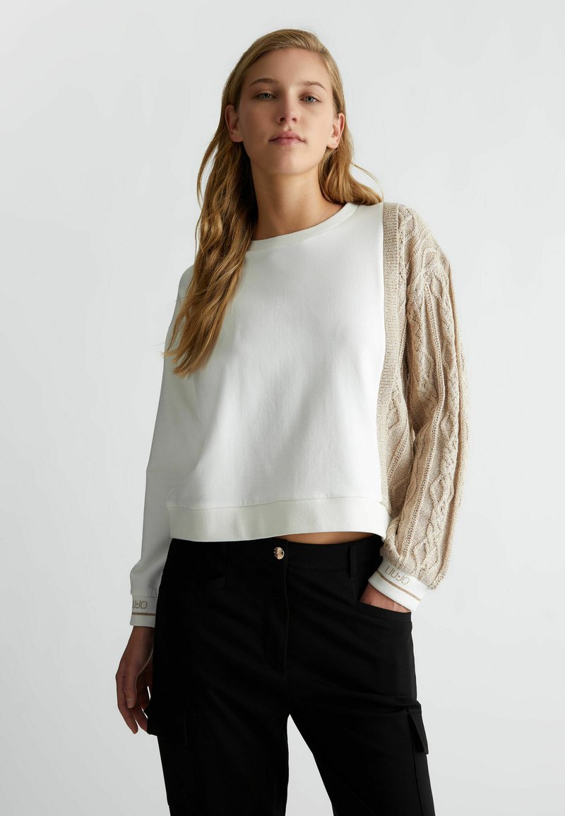 Felpa bianca corta con manica in maglia a treccia beige testurizzata, dotata di scollo rotondo e orlo a coste, abbinata a pantaloni neri.