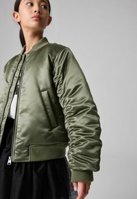 Giacca bomber in satin verde oliva con zip frontale, polsini a coste e una tasca laterale, abbinata a una gonna in tulle nera.