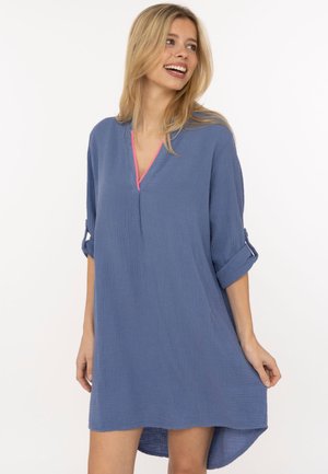 Blonde Frau lächelt, trägt ein lockeres blaues V-Ausschnitt-Kleid mit hochgekrempelten Ärmeln und rosa Besatz, steht vor weißem Hintergrund.