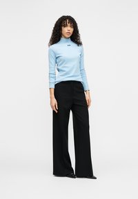 Lys blå turtleneck-genser med liten logo, kombinert med høytlivs svarte bukser med vide ben. Modell står foran en hvit bakgrunn.