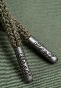 Lazos trenzados de color verde olivo con puntas de metal. Las puntas son plateadas con detalles grabados. La textura contrasta entre el metal suave y la tela tejida.