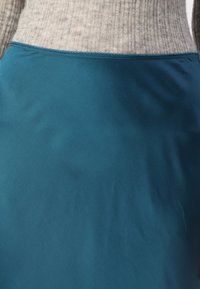 Teal satengskjørt med en glatt tekstur, med en kontrasterende sydd midjebånd i en lys nyanse. Enkel, tettsittende design.