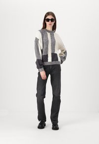 Gruby sweter dziergany w kolorze kremowym i szarym z wzorami warkoczy, połączony z czarnymi dżinsami o prostym kroju i czarnymi butami. Model nosi czarne okulary przeciwsłoneczne.