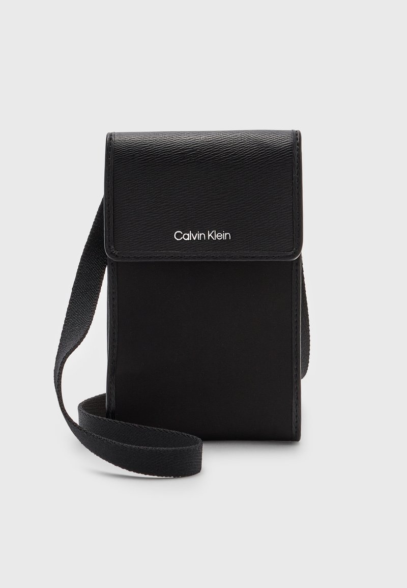Pochette noire Calvin Klein bandoulière avec fermeture à rabat et sangle en tissu ajustable, fabriquée en matériaux texturés et lisses.