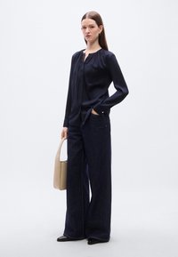 Μπλούζα από μετάξι σε ναυτικό μπλε με λαιμόκοψη-κλειδί, συνδυασμένη με φαρδιά τζιν παντελόνια σκουρόχρωμου denim. Το μοντέλο κρατά μία μπεζ τσάντα και φοράει μαύρα παπούτσια με μυτερό μπροστινό μέρος.