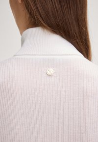 Weißer gerippter Pullover mit hohem Kragen und einem metallischen Logo-Akzent in der Mitte des Rückens. Die Textur wirkt weich und leicht.