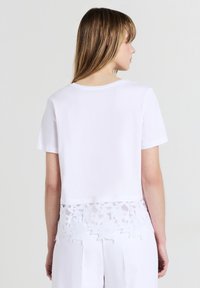 Donna con capelli biondi lisci, che indossa un crop top bianco a maniche corte con orlo in pizzo floreale e pantaloni bianchi, rivolta di spalle.