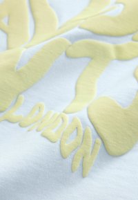 Světle modrý textil s vyvýšeným žlutým písmem. Text má hladkou texturu, plynulé tvary a výrazně zobrazené slovo "LONDÝN".