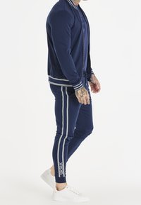 Mężczyzna ubrany w granatowy dres z jasnymi paskami i białe trampki, stojący bokiem na białym tle.