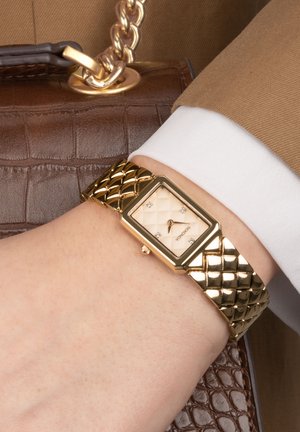 Montre-bracelet dorée avec un bracelet matelassé, un cadran rectangulaire, un fond crème et quatre accents scintillants, positionnée sur un poignet à côté d'un sac à main marron.