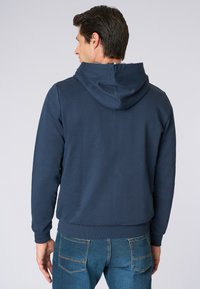 Felpa blu navy con cappuccio regolabile e polsini a coste. Texture liscia, design aderente, indossata con jeans blu.