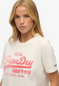 Biała bawełniana koszulka z dużym różowym nadrukiem, na którym widnieje napis "REAL SuperDry", oraz małą metką na lewym rękawie.