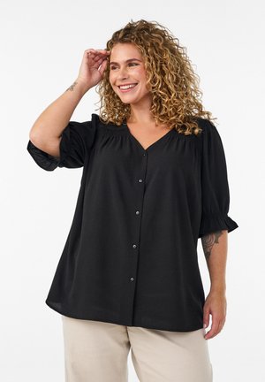 Žena s kovrčavom kosom koja se smiješi, nosi široku crnu košulju na kopčanje s nabranim rukavima do laktova i bež hlače na bijeloj pozadini.