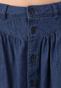 Denimhame korkean vyötärön kanssa, jossa on neljä mustaa nappia, laskostettu muotoilu ja tummansininen väri. Huomattavat tikkausvasteet näkyvissä.