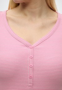 Κοντινό πλάνο ενός ριγέ πουκάμισου σε ροζ και λευκό χρώμα με V-λαιμόκοψη και τέσσερα ροζ κουμπιά μπροστά, που φοριέται από ένα άτομο με ανοιχτόχρωμο δέρμα και ξανθά μαλλιά.