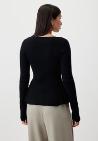 Black ribbed sweater with long sleeves, fitted body, and flared hem, worn over light-colored trousers. Smooth texture and minimalist design.