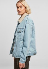 Svetlo modra jeans jakna z mekim bež šerpa ovratnikom, žepi na prsih in gumbi za zapiranje. Ohlapen kroj, vidni šivi.