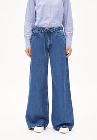 Jeans in denim a gamba larga di un blu medio, con una cintura regolabile con cordino, due tasche frontali e una texture scolorita. Abbinati a una camicia a righe.