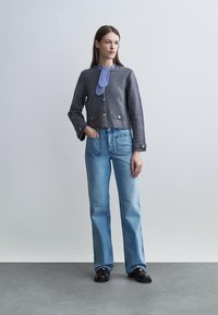 Femme debout portant une veste grise boutonnée, une cravate à rayures bleues, un jean bleu clair avec de grandes poches avant, et des mocassins noirs.