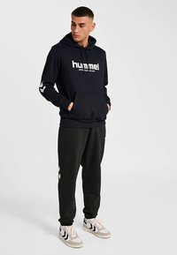 Svart luvtröja med framficka och vitt logo. Matchad med svarta byxor med vita ränder på sidorna och sneakers.