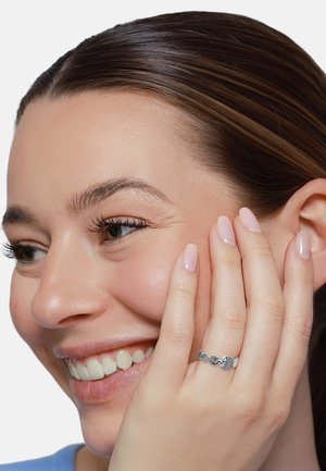 Lächelnde Frau mit braunen Haaren hält die Wange, zeigt einen silbernen Ring mit Herz und dekorativen Formen an ihrem Finger.