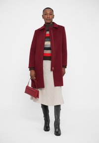 Weinroter Wollmantel mit Kragen und Reißverschluss, getragen über einem gestreiften Pullover, beigem Rock und schwarzen Kniestrümpfen. Beinhaltet eine kleine rote Handtasche.