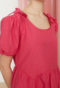 Roze jurk met pofmouwen en een ronde halslijn. Voorzien van strikken op de schoudernaden en een geplooide taille. Zacht materiaal met een gladde textuur.