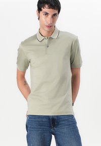 Šviesiai alyvuogių spalvos polo marškiniai su trimis mygtukais, kontrastingomis apykaklės juostelėmis raudonos, baltos ir juodos spalvų, bei mažas logotipo detalė.