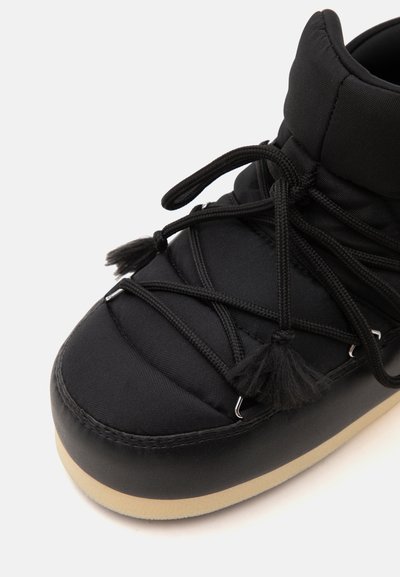Botte d'hiver noire matelassée avec semelle beige épaisse et lacets noirs croisés noués en pampilles décoratives sur fond clair.