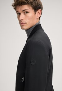 Jonge man in een zwart zip-up jasje met een merkpatch op de mouw en een grijze coltrui eronder, kijkend in de zijlijn.