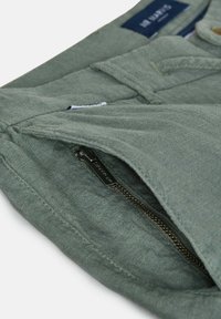 Pantalon en tissu vert avec une poche zippée partiellement ouverte montrant une tirette en métal et une étiquette de marque visible sur la ceinture.