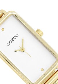 Montre rectangulaire dorée avec un cadran blanc, trois aiguilles dorées et un logo discret. Elle possède un bracelet en or rainuré et une couronne texturée.