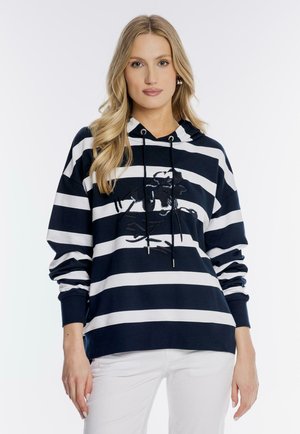 Blondynka w granatowym i białym bluzie z poziomymi paskami i czarnym haftowanym wzorem, w białych spodniach, na białym tle.