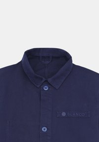 Chemise bleu marine avec col boutonné, dotée de deux boutons bleus, et d'un patch texturé portant l'inscription « BLANCO » sur le côté gauche.
