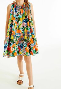 Bunt gemustertes Blumenkleid mit Rüschenausschnitt, gestuftem Design und lebendigen Mustern. Kombiniert mit weißen Riemchensandalen.