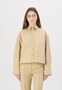 Beige cropped shirt with a collar, front buttons, and a chest pocket. Features wide sleeves and embroidered logo detail.