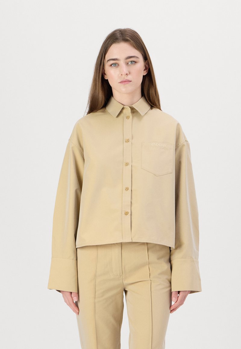 Beige cropped shirt with a collar, front buttons, and a chest pocket. Features wide sleeves and embroidered logo detail.