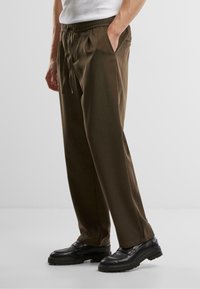 Pantalons marron à taille élastique avec poches latérales, jambes fuselées et texture lisse, accompagnés de mocassins noirs épais.