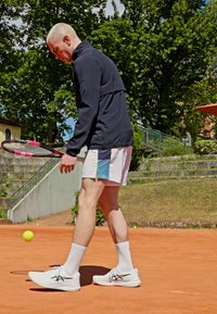 Sportiv masculin îmbrăcat într-o jachetă neagră și șorturi multicolore, ținând o rachetă de tenis, mergând pe un teren de zgură cu o minge de tennis aproape.
