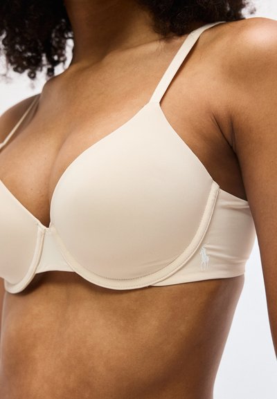 Μπεζ σουτιέν με μπανέλα και λείες κύπελλες, φορεμένο από άτομο με μεσαίο τόνο δέρματος και κατσαρά μαλλιά, με ορατό κεντημένο λογότυπο στη ζώνη.