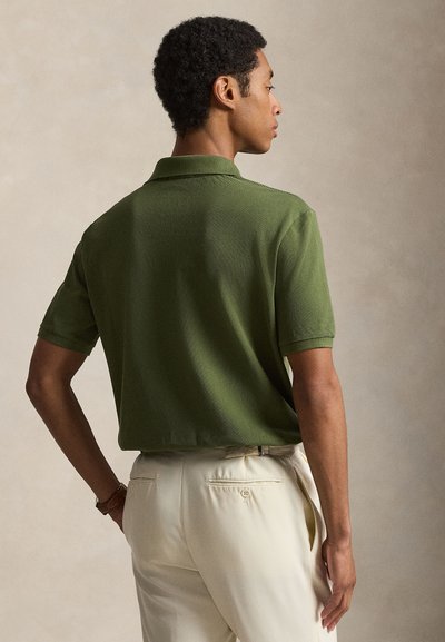 Άνδρας που κοιτάζει πλάγια και προς τα πίσω, φορώντας πουκάμισο πόλο με κοντά μανίκια σε ελιά πράσινο χρώμα και ανοιχτό μπεζ παντελόνι, μπροστά σε ένα απλό φόντο.