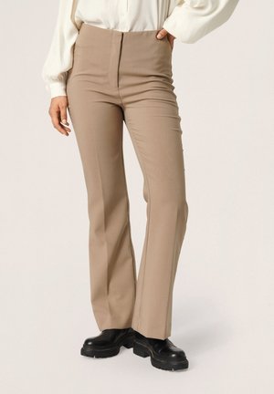 Vrouw draagt beige rechte broek, zwarte stevige laarzen en een wit blouse met lange mouwen, staande met één hand in de zak.