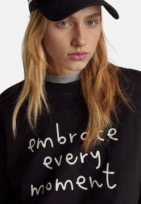 Sudadera negra con texto blanco "abraza cada momento". Cuello alto gris visible por debajo. El modelo lleva una gorra negra.