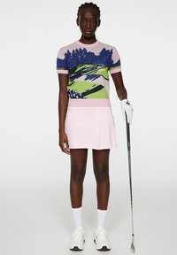 Sweter golfowy dzianinowy w pastelowym różowym kolorze, z graficznym motywem niebieskiego i zielonego krajobrazu. Połączony z jasnoróżową spódnicą plisowaną. Białe rękawiczki i buty.