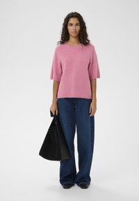 Kurzärmeliges, hellrosa gestricktes Sweatshirt mit rundem Ausschnitt, kombiniert mit dunkelblauen Jeans mit weitem Bein und einer schwarzen Tasche. Durchgehend glatte Textur.