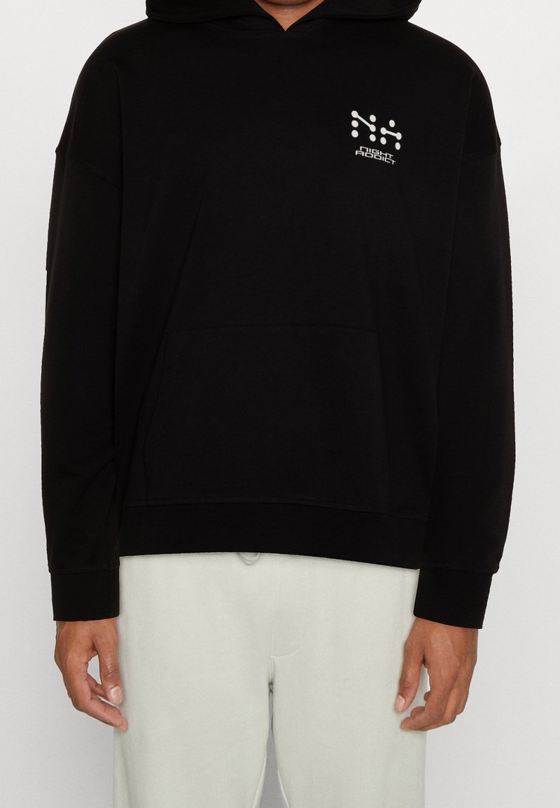 Personne portant un sweat à capuche noir avec le logo blanc « Night Addict » sur la poitrine gauche et un pantalon clair, debout devant un fond uni.